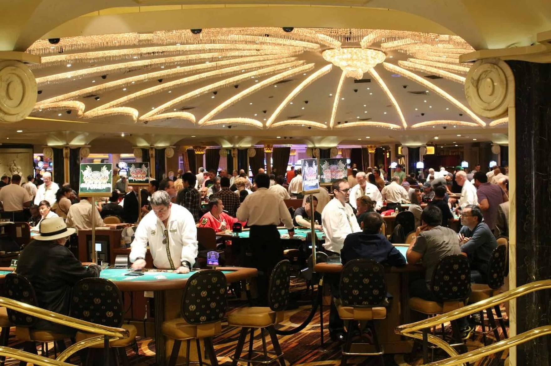 A busy casino table games pit, dealers and players at card tables under an ornate chandelier ceiling.